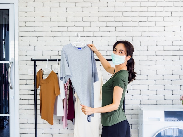 Young beautiful woman, housewife in casual wearing protective face mask hanging dry shirt with hanger on clothesline after washing near wash machine in laundry room on white wall. Premium Photo